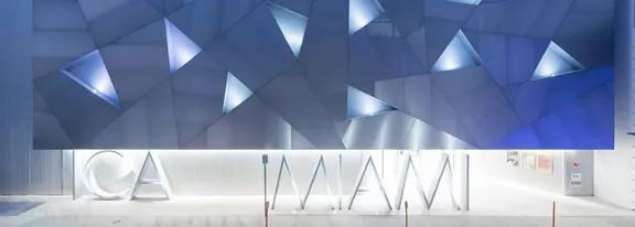 A contemporary building facade features a geometric pattern of triangular panels illuminated in cool blue and white light. Large sculptural letters spelling out 'ICA MIAMI' stand prominently at the entrance. The scene appears to be captured at night or dusk, emphasizing the glowing architectural details. The overall atmosphere is sleek, minimalist, and futuristic.
