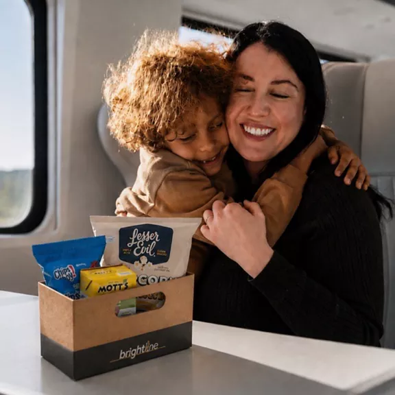A smiling woman and young child share a warm hug while seated on a modern train. In front of them is a cardboard snack box with the word "brightline" clearly visible on the side. Assorted snack packages are arranged inside the box, adding color and detail to the scene. Sunlight streams through the window, creating a cozy and relaxed travel atmosphere.