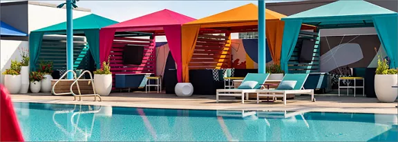 A row of vibrant poolside cabanas in pink, orange, teal, and blue fabric lines the deck beside a clear swimming pool. White lounge chairs and small tables sit under the shade structures, creating a relaxed resort atmosphere. Large potted plants and modern architectural elements frame the scene, while reflections shimmer on the calm water. The image conveys a bright, sunny day with a contemporary, tropical style.