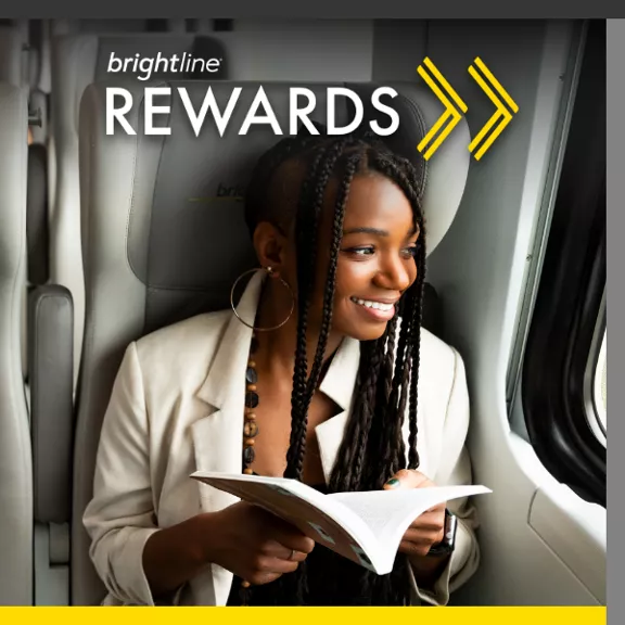 A young woman sits on a Brightline train, smiling while reading a book. The text "brightline REWARDS" appears prominently at the top with two yellow arrows pointing right.