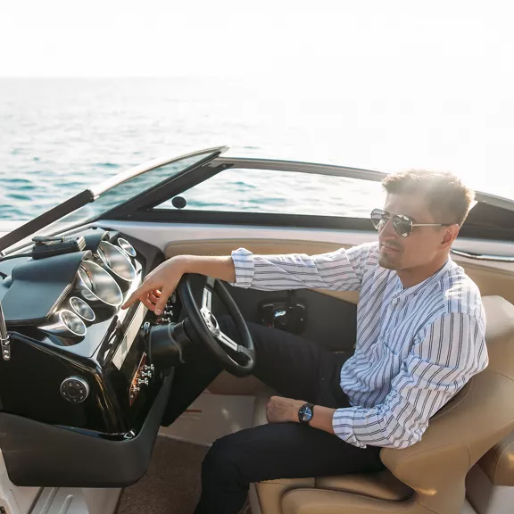 A well-dressed man sitting at the wheel of a boat.