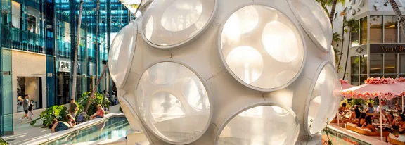 A large white geometric sphere sculpture stands beside a narrow reflecting pool in an outdoor shopping plaza. Modern glass-fronted buildings and storefronts surround the artwork, with palm trees adding a tropical atmosphere. People sit and walk under pink umbrellas at a café area in the background. Warm sunlight creates bright reflections on the sculpture and nearby architecture, giving the scene a vibrant, contemporary feel.