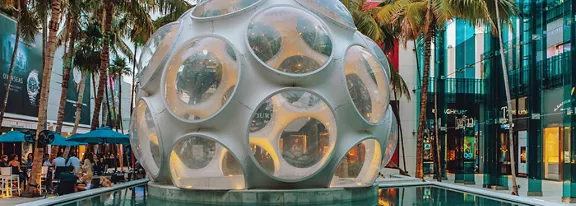 A large spherical glass and metal structure with circular windows stands at the center of an outdoor plaza. The sculpture is surrounded by a shallow reflective pool and framed by tall palm trees. Modern glass-fronted buildings and outdoor café seating with umbrellas create an urban lifestyle setting. Warm lighting and evening tones give the scene a vibrant, contemporary atmosphere.
