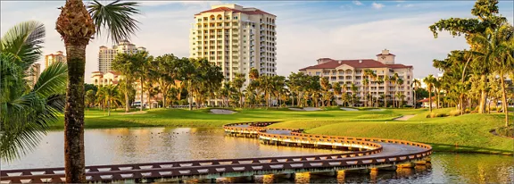 A scenic lakeside resort features tall condominium-style buildings surrounded by lush palm trees and manicured lawns. A curving wooden boardwalk crosses the water, leading the eye toward the waterfront architecture. The scene is bathed in warm golden light, suggesting late afternoon or sunset. Calm water, greenery, and clear skies create a tranquil, vacation-like atmosphere.