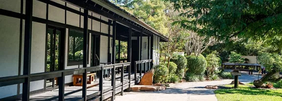 A long, modern building with Japanese-style architectural elements stands beside a landscaped garden. Large windows and dark wooden beams frame a shaded walkway with simple benches. Lush green trees, shrubs, and rocks surround the structure, creating a peaceful outdoor environment. Sunlight filters through the foliage, highlighting the tranquil, natural atmosphere.