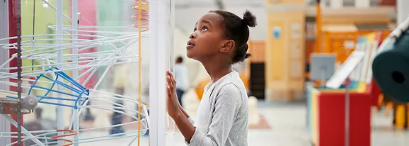 A young girl stands closely in front of a transparent interactive exhibit, gazing upward with curiosity. The setting appears to be a bright indoor science center or museum with colorful structures in the background. Tubes and wires inside the display suggest a hands-on educational installation. The overall scene conveys exploration, learning, and wonder in a playful environment.