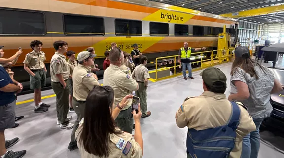 A group of scouts and adults gather inside a large maintenance facility beside a Brightline passenger train. The train features a yellow and orange gradient design with the words brightline and SMART SERVICE visible on the side. A staff member in a safety vest addresses the group from a platform next to the train.