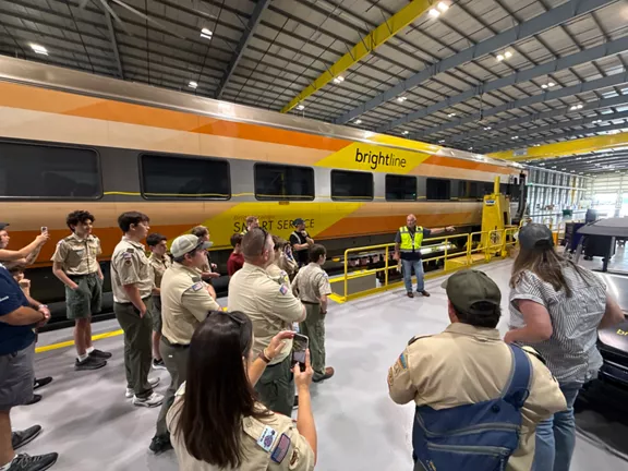 A group of scouts and adults gather inside a large maintenance facility beside a Brightline passenger train. The train features a yellow and orange gradient design with the words brightline and SMART SERVICE visible on the side. A staff member in a safety vest addresses the group from a platform next to the train.