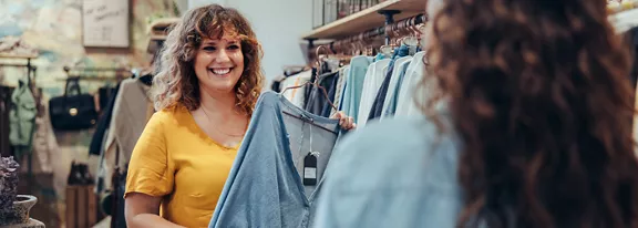 A cheerful woman in a yellow top holds up a light denim shirt on a hanger inside a clothing store. She appears to be showing the shirt to another shopper, creating a friendly and social shopping moment. Racks of clothes and hangers fill the background, suggesting a boutique retail environment. The overall scene feels casual, warm, and inviting.