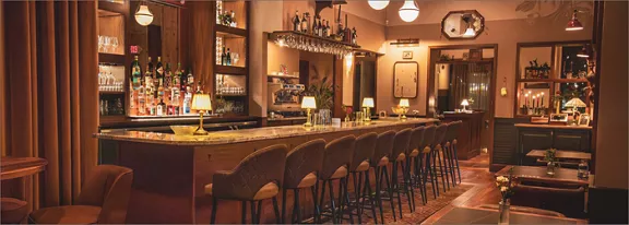 A stylish bar interior features a long marble-topped counter lined with upholstered bar stools. Shel
