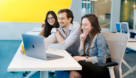 Three smiling Brightline employees, sitting at a table working on a silver mac computer