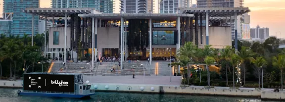 A contemporary museum building stands along a waterfront promenade lined with palm trees. Wide steps lead up to the entrance, where people gather under a large overhanging roof structure. In the foreground, a boat with a large digital screen cruises along the water, displaying illuminated text and graphics. The city skyline with high-rise buildings rises in the background at sunset, casting a warm glow over the scene.