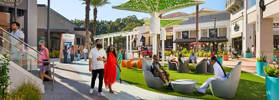 An open-air shopping center features people walking and relaxing on contemporary seating under sculptural shade structures. The scene includes retail storefronts, palm trees, and landscaped planters surrounding a central lawn area. Shoppers and visitors of various ages are engaged in casual conversation, leisure, and window shopping in bright daylight. The overall atmosphere is vibrant, social, and inviting, highlighting a modern urban lifestyle destination.