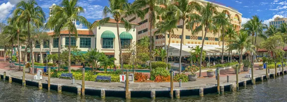 A vibrant waterfront promenade features a row of palm trees lining the edge of a calm canal or river. Colorful low-rise buildings with awnings and balconies house shops, restaurants, or cafes. Benches, planters, and landscaped greenery create a welcoming pedestrian area along the water. The scene suggests a warm, tropical climate under a bright blue sky with scattered clouds.