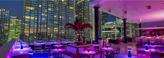 A modern rooftop bar is illuminated with vivid purple and blue lighting against a nighttime city skyline. Tall glass buildings with many lit windows surround the open-air terrace, creating an urban atmosphere. Tables and seating are arranged around a central planter with a palm tree, and the bar area is visible to the right. The word "CHASE" appears brightly on one of the nearby buildings, adding a recognizable city landmark to the scene.