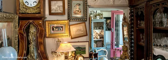 The image showcases a richly decorated antique shop filled with ornate furniture, framed artwork, and glassware. A large clock with Roman numerals is prominently displayed, surrounded by gilded frames and classic lamps. The setting is cozy and nostalgic, featuring a mix of warm wood tones and pastel accents. Reflections and mirrors add depth, while the overall mood is inviting and timeless.