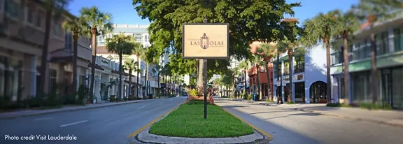 A wide view of Las Olas Boulevard shows a sunlit, empty street lined with palm trees and colorful storefronts. The central focus is a sign reading 'Las Olas' with a decorative emblem, positioned on a grassy median. The scene is bright and inviting, with clear blue skies and a relaxed urban atmosphere. The image includes the visible text 'Las Olas' and 'Photo credit: Visit Lauderdale' in the lower left corner.