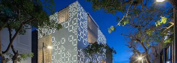 A contemporary building features a striking geometric facade, illuminated with patterned lights during twilight. The structure stands out against a deep blue evening sky, surrounded by leafy trees and urban streetlights. The scene captures a blend of modern architecture and natural elements, with no visible text or numbers present. The overall mood is calm and sophisticated, highlighting urban design.