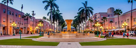 The image features a vibrant outdoor plaza with a large central fountain surrounded by palm trees and pastel-colored buildings. People are seen walking and gathering, creating a lively atmosphere. The setting is illuminated by ornate street lamps, and the sky displays a soft blue and pink gradient, enhancing the relaxed mood. No visible text or numeric values are present in the image.