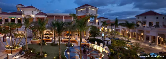 The image features a spacious, upscale outdoor shopping plaza surrounded by palm trees and elegant Mediterranean-style architecture. The setting is captured at dusk, with soft lighting illuminating walkways and landscaped gardens. People are visible enjoying the area, creating a lively yet relaxed atmosphere. The sky is partly cloudy, adding depth to the evening ambiance. No explicit numbers or identifiable brands are visible.