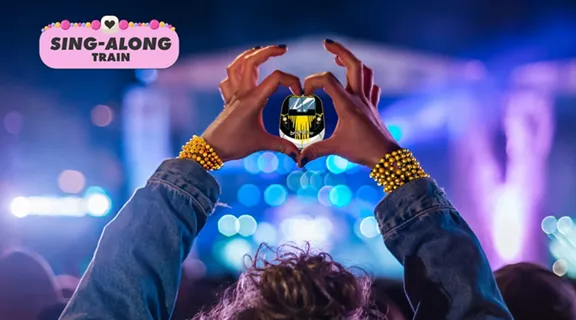 A young lady making a heart shape with her hands and fingers at a concert with an image of a Brightline train inside the heart and a graphic above that reads sing-along-train