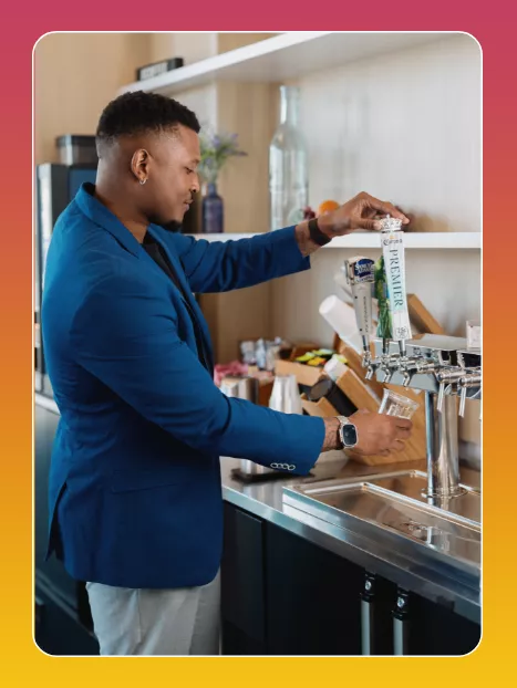 A man in a blue blazer pours a drink from a draft tap into a clear plastic cup. He stands at a sleek counter in a contemporary bar or office refreshment area. 