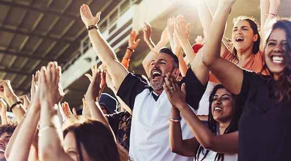 Crowd of sports fans cheering during a match in stadium. Excited people standing with their arms raised, clapping, and yelling to encourage their team to win.