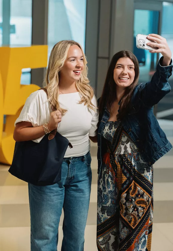 Two young women stand together in a Brightline station lobby while one holds up a smartphone to take a selfie. 