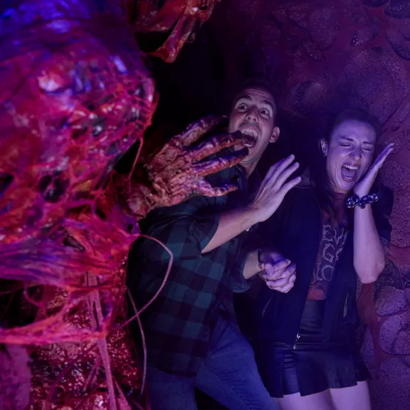 A monstrous, red, sinewy creature reaches toward a frightened couple in a dark, cavernous setting. The man and woman cling to each other, screaming and recoiling in fear from the approaching horror.