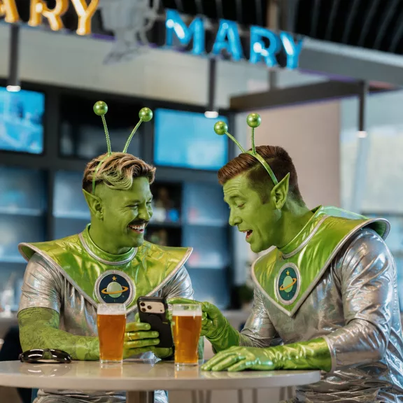 Two men dressed as green aliens sit at a bar table, smiling and looking at a smartphone together. They wear shiny silver and green space-themed costumes with antenna headbands and planet emblems on their chests. Two pints of beer are placed on the table in front of them.