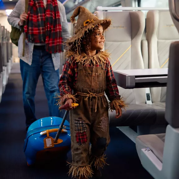 A young child dressed in a scarecrow costume walks down the aisle of a modern passenger train. The child wears a straw-trimmed hat, patched overalls, and a plaid shirt, pulling a bright blue rolling suitcase. 