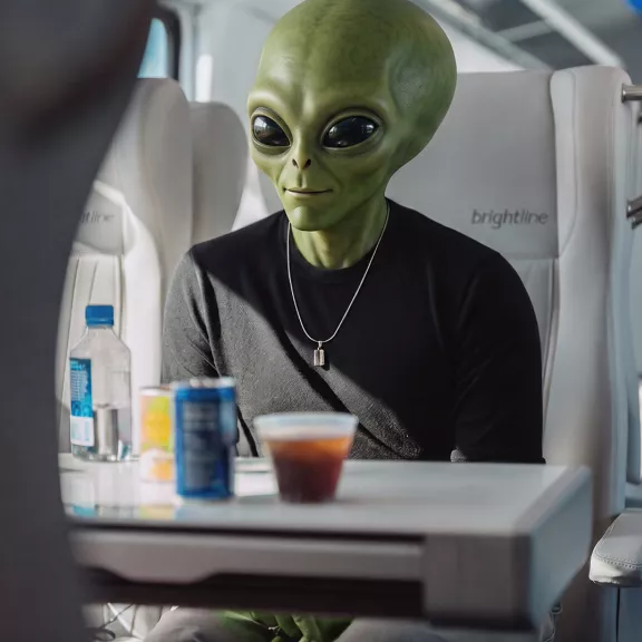 A green alien figure sits calmly in a bright, modern train seat with the word "brightline" visible on the headrest. The character wears a black shirt and a necklace.