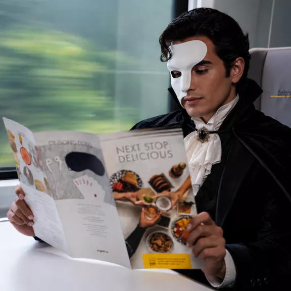 A young man wearing a white half mask and a dramatic black cape sits on a modern train, reading a colorful food brochure. The brochure cover features the headline "NEXT STOP: DELICIOUS" above an assortment of dishes. 