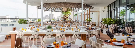 Contemporary open-air rooftop restaurant featuring white tables and sculptural chairs overlooks a distant city skyline. A central bar with a thatched-style canopy anchors the space, surrounded by neatly arranged place settings and amber glassware. Potted greenery and cushioned lounge seating add warmth and comfort to the bright, airy terrace. The overall scene suggests a relaxed yet upscale dining experience in an urban setting.
