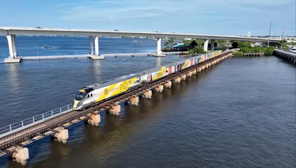 A sleek yellow and white passenger train travels along a low rail bridge over a wide body of water. The train features colorful graphics on its carriages as it moves parallel to a higher concrete highway bridge. The scene is set under a clear blue sky with calm water and distant shoreline structures. The image conveys modern transportation and coastal infrastructure in a bright, sunny environment.