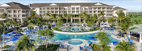 A large luxury resort hotel is shown with multiple stories and balconies overlooking an expansive pool complex. The scene features a central curved swimming pool, a round hot tub, and numerous lounge chairs with blue umbrellas. Palm trees and lush greenery surround the water areas, creating a tropical atmosphere. The background reveals open green fields beyond the resort, suggesting a spacious, tranquil vacation setting.