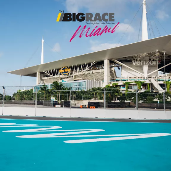 A vibrant race track is pictured in front of a large stadium, with clear signage for 'The Big Race Miami' and 'verizon' visible. The setting is outdoors under a bright sky, with palm trees and the stadium structure dominating the background.