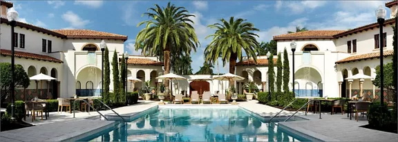 A large outdoor swimming pool sits at the center of a Mediterranean-style resort courtyard. Lounge chairs and umbrellas line both sides of the pool, creating a relaxed vacation atmosphere. Tall palm trees and manicured shrubs frame the scene, with stucco buildings and terracotta roofs in the background. The clear blue sky and reflections in the water emphasize a sunny, tranquil setting ideal for leisure and relaxation.