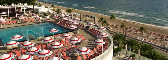 A luxurious beachfront resort features a large outdoor swimming pool surrounded by rows of red and white umbrellas and sun loungers. The scene overlooks a sandy beach lined with palm trees and the ocean waves in the background. The elevated terrace area creates a panoramic view of the coastline and leisure facilities. The overall atmosphere suggests a sunny, relaxing vacation destination.
