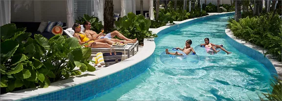 A family relaxes at a resort beside a winding lazy river pool. Two adults lounge on chairs under a s