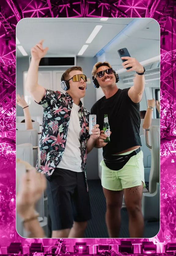 Two men enjoying drinks on a Brightline train while taking a selfie.