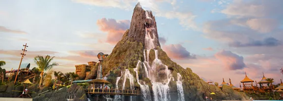 A towering artificial volcano with cascading waterfalls dominates a tropical water park scene. Woode