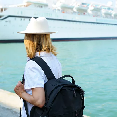 A young lady wearing a white fedora hat and carrying a black backpack walking along the water in a seaport looking at a large cruise ship in the distance.