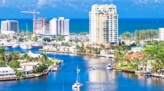Panoramic view of a modern coastal city with high-rise buildings along a blue waterfront. A wide canal or river runs through the scene, bordered by lush green palm trees and residential areas. The distant ocean horizon and clear sky suggest a bright, sunny day. Construction cranes and varied architecture highlight ongoing urban development and a vibrant cityscape.