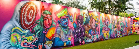 A long outdoor mural features a series of surreal, cartoon-like characters painted in vivid colors. The artwork includes human and animal hybrids, futuristic figures, and fantasy creatures rendered in neon tones. The mural is set in a landscaped urban area with green grass and palm trees in the background. Bright pinks, blues, and greens dominate the palette, creating a vibrant, energetic street art scene.