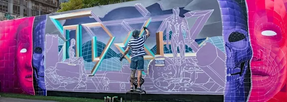 A street artist stands on a small platform, painting a large, colorful mural on an outdoor wall. The artwork features futuristic geometric shapes, outlined figures, and two large stylized faces on each side in vivid pink and purple tones. Spray paint cans are arranged on the ground in front of the mural, suggesting an ongoing creative process. Urban buildings and greenery are visible in the background, placing the scene in a city environment.