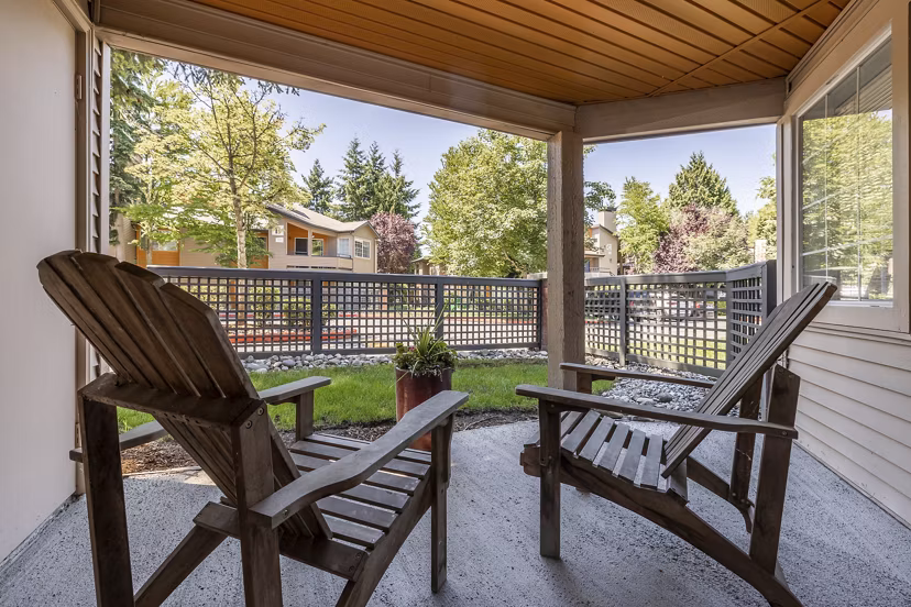 Cozy covered patio with two wooden chairs and a small table overlooking lush greenery and nearby buildings