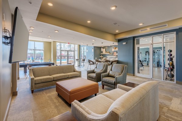 Modern lounge area with comfortable seating and large TV, overlooking a fitness room with exercise equipment
