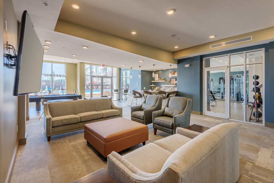 Modern lounge area with comfortable seating and large TV, overlooking a fitness room with exercise equipment