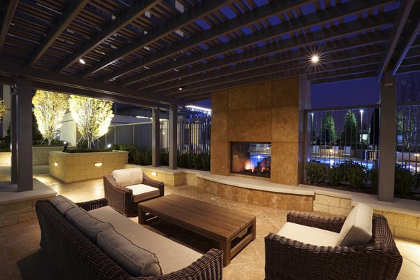 Outdoor patio with wicker furniture, wooden table, and fireplace surrounded by softly lit trees